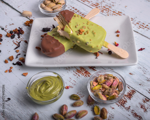 Artisanal pistachio ice cream bars dipped in chocolate served on a white plate with pistachio cream, shelled nuts, and star anise on wooden background