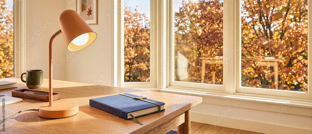 Naklejka premium Illuminated orange desk lamp and closed blue notebook on wooden table in cozy home office workspace. Large windows displaying bright golden autumn foliage and warm fall sunlight with copy space