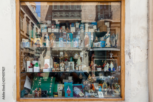 A religious souvenir shop window in Valletta displays statues, icons, and figurines, its glass reflecting the balconied street outside, Malta