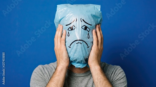 Person holding transparent plastic bag over head with drawn sad face against blue background with expression against solid featuring drawn everyday life person covers drawing face translucent with sky