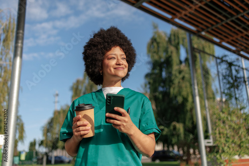 Smiling nurse in scrubs holding coffee and smartphone, enjoying a sunny break