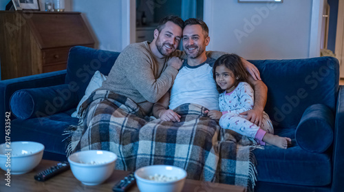 Cozy Evening Together: A happy family, including two fathers and their daughter, enjoy a movie night on the couch, sharing popcorn and warmth under a checkered blanket.