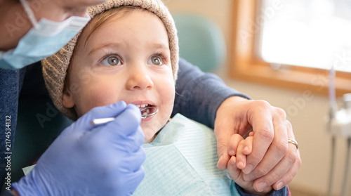 Young Patient's First Dental Visit: A young child, looking upwards with curiosity, undergoes a dental check-up, held gently by a parent. A dentist, wearing gloves and mask.