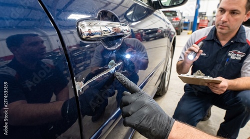 Professional car mechanic inspecting vehicle body damage with a clipboard in a modern auto repair shop garage