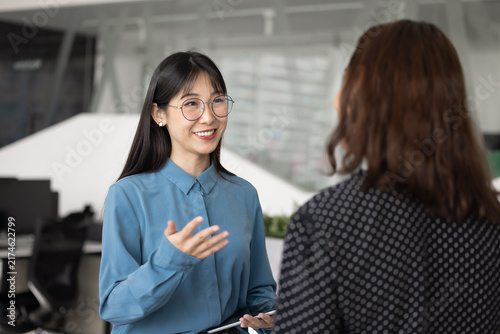 Young Asian professional woman having friendly discussion  informal conversation with female colleague standing in modern coworking space. Business communication  collaboration  workplace interaction