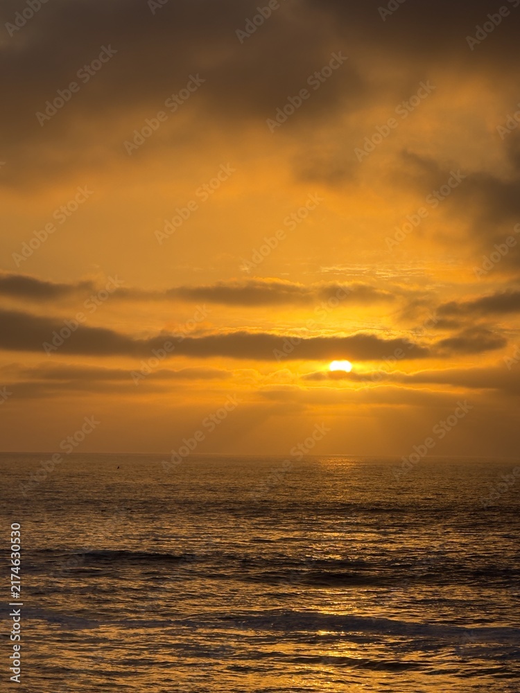 Fototapeta premium Vertical photography of intense golden orange sunset sun rays breaking through sky clouds over ocean in La Jolla, CA.