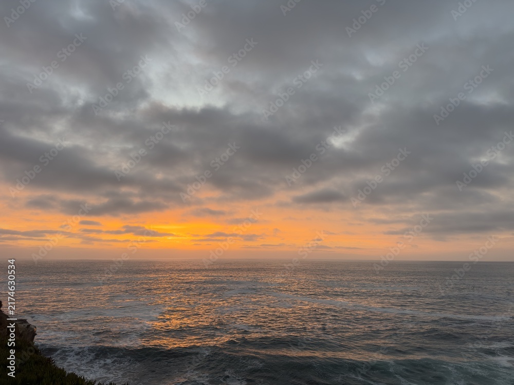 Fototapeta premium Wide vista of brilliant orange afterglow along the ocean horizon under dark overcast evening clouds in La Jolla, CA.