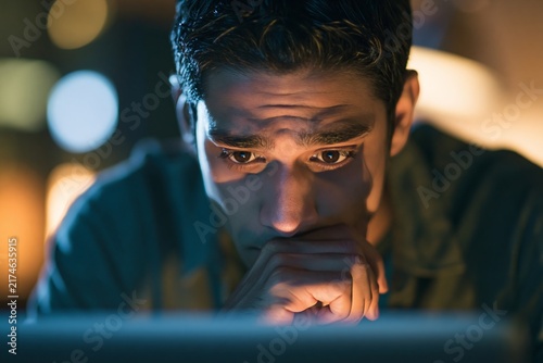 Close up of young man looking intently at a computer screen in a dark room at night with warm glow