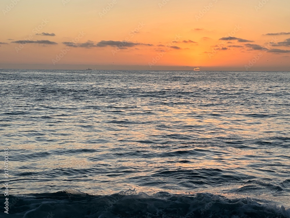 Obraz premium Vibrant orange sunset over calm ocean waters in Carlsbad, California with distant boat outline on horizon.