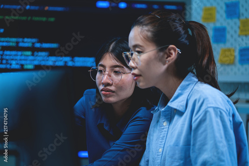 Asian technology professionals collaborating while reviewing computer code and digital information during software development work. The scene represents programming teamwork