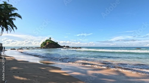 Wide daytime view of a Sri Lanka beach featuring a large rocky outcrop and breakwaters with the Indian Ocean in the background under bright sunlight.
