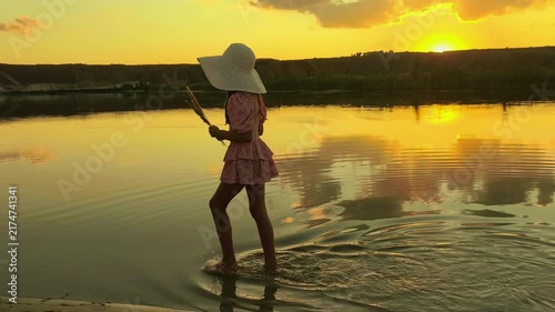 Young girl wearing a sun hat wades through shallow lake water during golden hour, peaceful childhood, summer travel and scenic outdoor lifestyle.