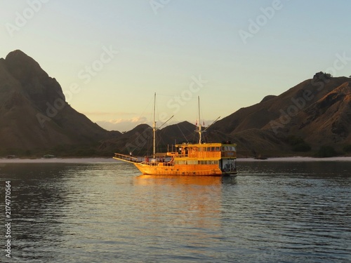 Fotografie Traditional wooden phinisi boat anchored at golden hour, Padar Island, Komodo, I