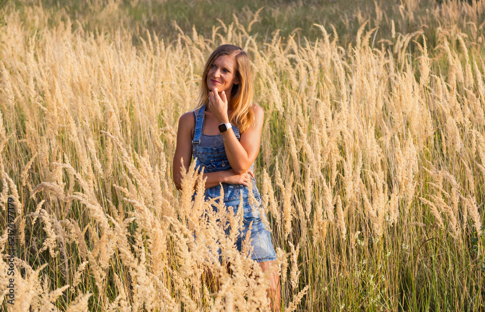Obraz premium Pensive attractive woman posing in high dry grass field at sunset, beautiful rustic lifestyle portrait.