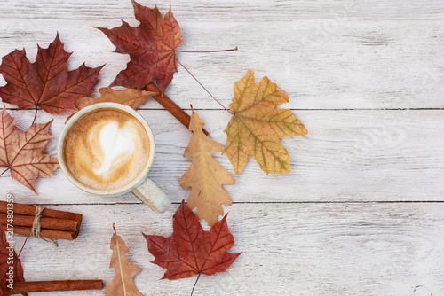 Фотография Mug of spicy coffee with milky foam, red maple leaves on white wooden table