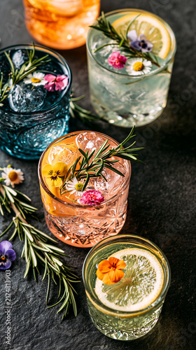 Colorful cocktail glasses garnished with rosemary, citrus and edible flowers arranged on a dark stone surface. Elegant botanical beverage display.