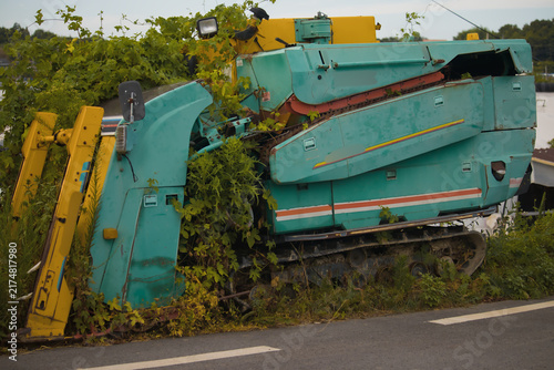 Quadro su tela Abandoned Teal and Yellow Industrial Machine Overgrown with Green Vines by an As