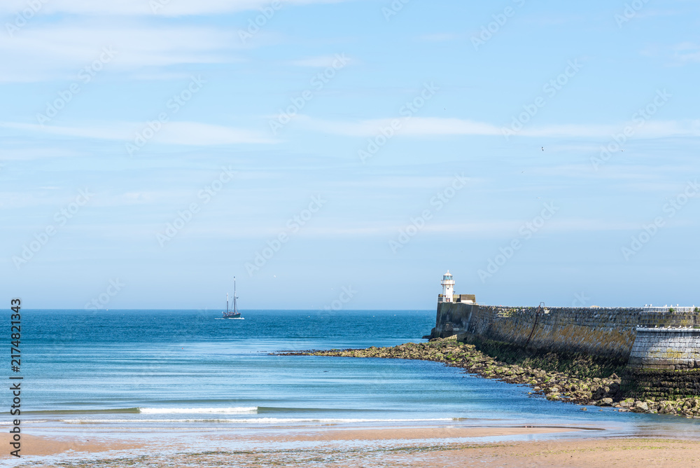 Fototapeta premium A calm North Sea scene along the Scottish coast, featuring a lighthouse with a green navigation light on a stone pier, a distant sailing vessel and gentle waves reaching the sandy shoreline under clea