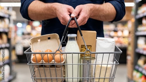 Close-up of a male customer's hands holding a full grocery basket in front of them in a supermarket. Retail daily, food shopping and consumer lifestyle concept