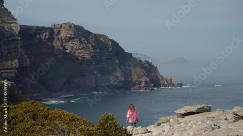 Young woman hikes along Cape of Good Hope cliffs, pausing above blue ocean as rugged coastal mountains, rocky shoreline, and low shrubs frame scenic outdoor travel adventure in daylight.