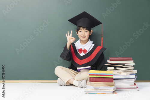 Foto School child in graduation cap with books making OK sign by chalkboard