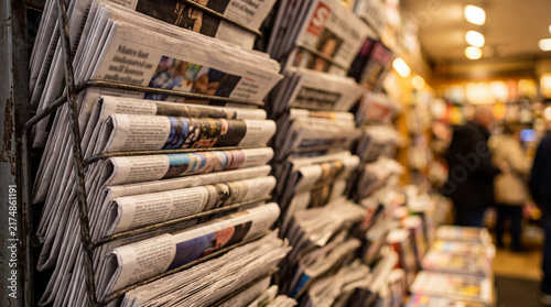Newspapers stacked in a store showing different sections and headlines in a busy location during the day