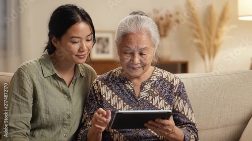 Asian Grandmother and Granddaughter Learning to Use Digital Tablet Together on Cozy Sofa
