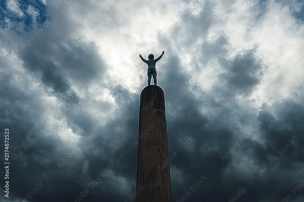 Obraz premium solitary human figure sculpture with arms raised atop a tall cylindrical column against dramatic stormy clouds, moody triumphant and defiant atmosphere