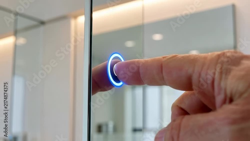 Finger pressing illuminated touch button on smart mirror during daytime with soft reflections