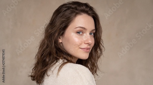 Young woman with wavy brown hair and blue eyes looking over her shoulder with a slight smile against a textured neutral background