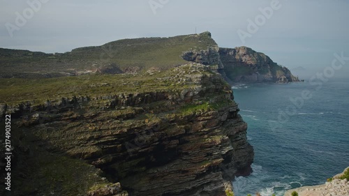Coastal cliffs rise above blue ocean waters, revealing rugged rock strata, green terrain and a distant headland beneath hazy sky. Natural seascape provides scenic space for travel and nature themes.