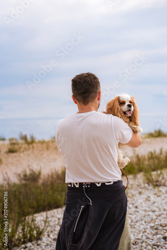 Quadro su tela Man seen from behind carrying a Cavalier King Charles Spaniel in his arms on a w