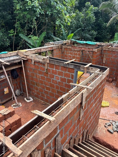 House construction site in Kerala, India, showing a partially built residential home with concrete columns, exposed brick walls, construction materials, and workers’ activity.