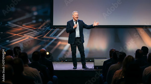 Middle aged man wearing business suit speaks into microphone while standing on stage before live audience during corporate conference event held inside auditorium venue with large screen