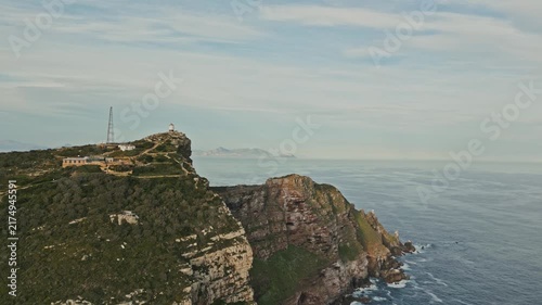 Rocky coastal cliffs rise above calm ocean water, with green vegetation, layered rock faces, and small hilltop structure beneath soft blue sky, creating serene natural landscape view from distant