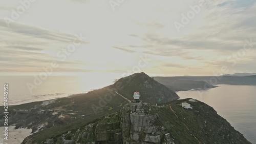 Aerial view of Cape of Good Hope coastline with lighthouse on rugged mountain cliffs above ocean, glowing in warm evening light beneath layered clouds and vast scenic horizon at sunset.