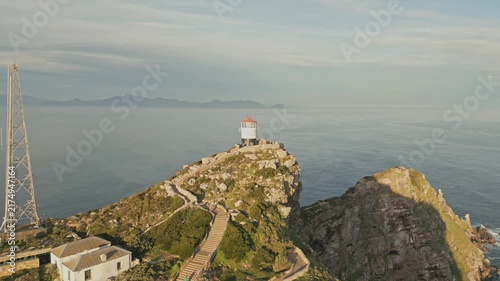 Drone pull back from white lighthouse on rocky sea cliff, revealing winding staircase, communications tower, coastal buildings, calm open water, distant islands, and warm sunlight under thin clouds.