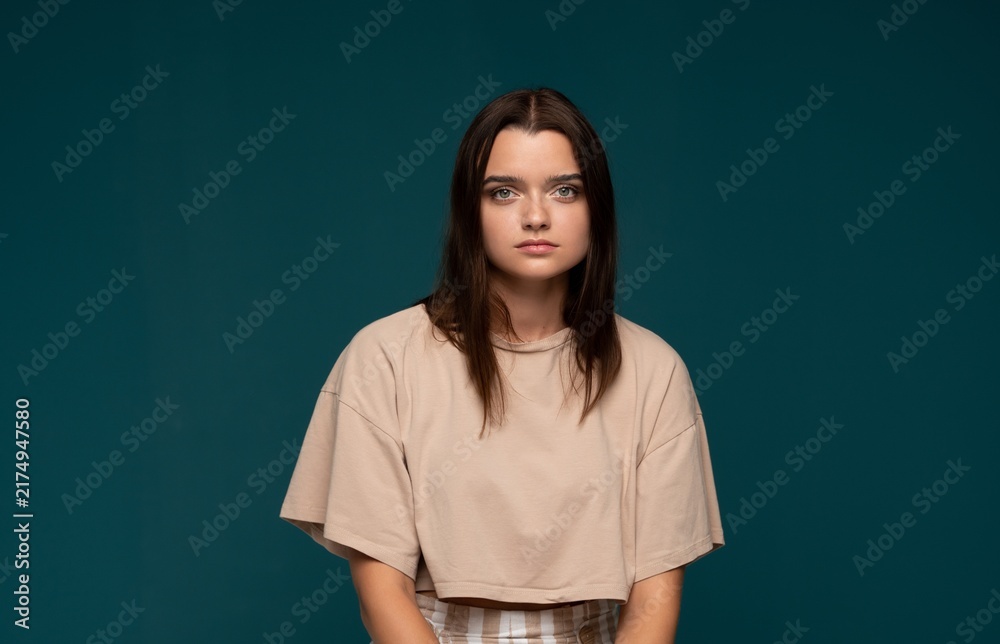 Obraz premium Young woman with neutral expression posing, wearing casual crop top against a teal background