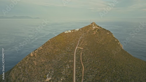 Drone aerial view of lighthouse atop green mountain ridge above calm sea, with narrow summit trail, rocky slopes, hazy horizon, and distant islands under soft daylight in serene setting.