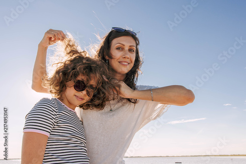 smiling mother gently touching daughter hair while both looking at camera outdoors under bright blue sky by the water with natural sunlight and relaxed family moment, copy space, family travel,