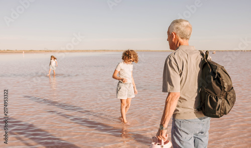 senior man in shallow pink lake watching children exploring water, family outing at sunset. peaceful outdoor travel with backpack, family travel tourism, multigenerational bonding.