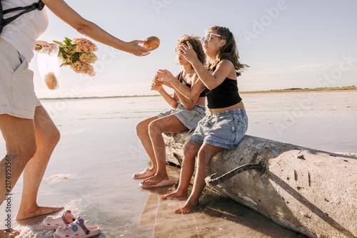 mother handing pastry to children sitting on driftwood log at shallow salt lake, sunny family outing with warm summer light and joyful vacation atmosphere, family travel, tourism, bakery