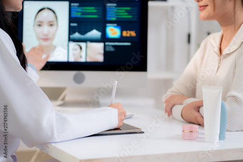 Asian woman doctor writing medical note on tablet for woman patient while computer screen with ai skin analysis and digital scan result, ai technology data for cosmetic aesthetic, consultation beauty.