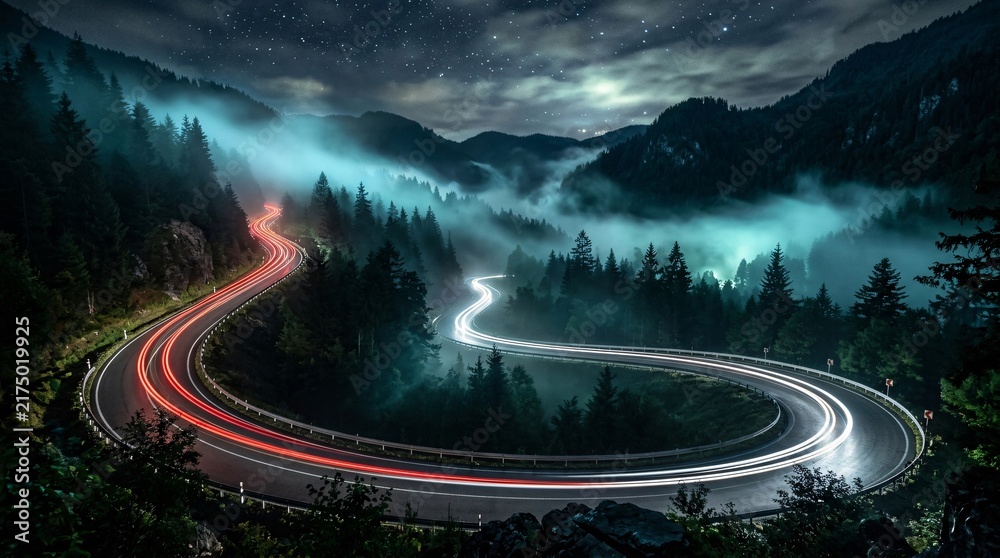 Obraz premium Winding mountain highway at night in thick glowing fog, long exposure car light trails on curved asphalt, eerie cinematic scenery.