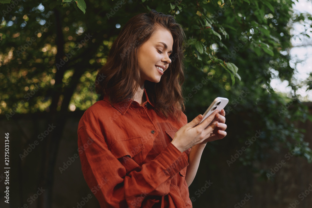 Naklejka premium Woman lifestyle shot outdoors wearing a red shirt, smiling while using a smartphone, natural light, green trees in the background, relaxed candid moment in a park or garden setting