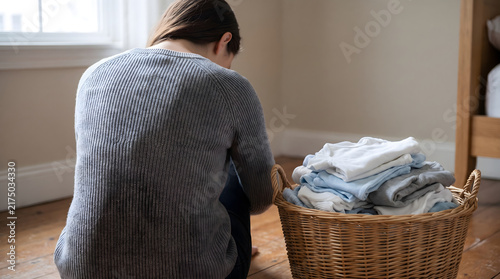 Tired mother burying face in arms beside laundry basket of folded baby clothes dealing with postpartum anxiety