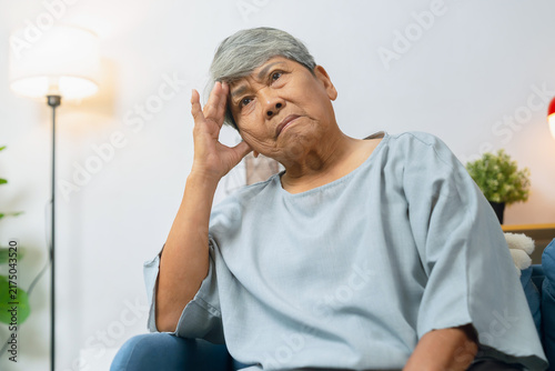 Stressed elderly Asian woman sitting on a sofa at home with a headache, experiencing fatigue and emotional stress.