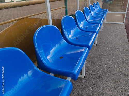 Outdoor sports dugout featuring bright blue plastic seats arranged beneath a clear canopy for players, coaches, and substitutes.