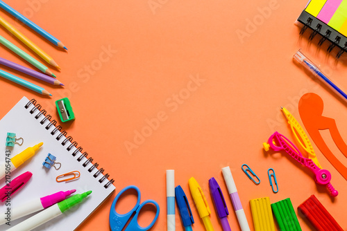 Creative flat lay of colorful pencils, markers, scissors, notebook, clips and stationery arranged around an orange background with copy space.