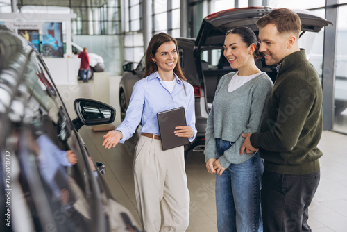 Salesperson assisting young couple at car dealership, explaining features of new automobile in modern showroom